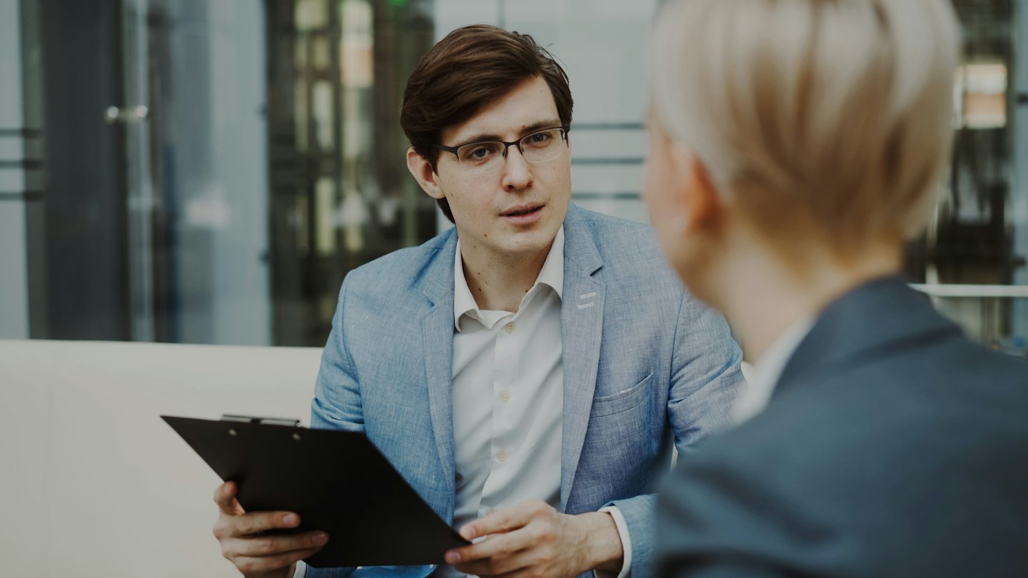 Man in glasses holding a clipboard and speaking to someone.